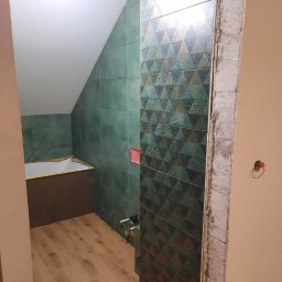 LUX-HAUS - Wnętrze łazienki w trakcie prac wykończeniowych, widoczne zielone płytki na ścianach, wzorzysta ściana z płytek, drewnopodobna podłoga oraz fragment wanny w skosie pomieszczenia.
