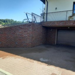 LUX-HAUS - Ceglana elewacja budynku jednorodzinnego z garażem, widoczny balkon z metalową balustradą i pies na nim, teren wokół wyłożony kostką brukową