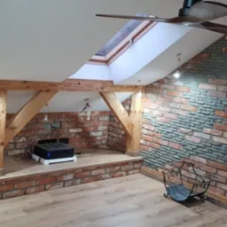 Poddasze z ceglaną ścianą, drewnianymi belkami, oknem dachowym i wentylatorem sufitowym, częściowo wykończone szarą okładziną imitującą kamień.