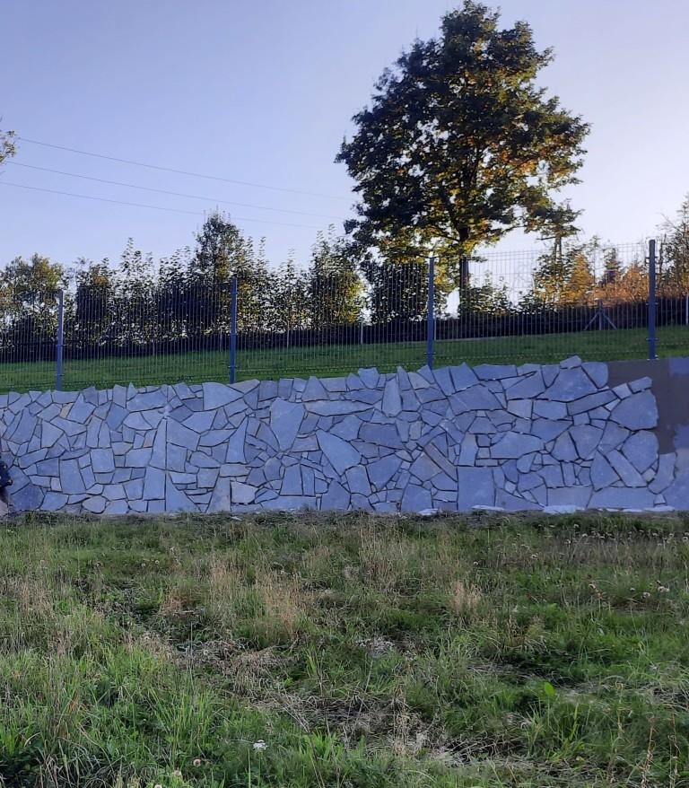 Mur oporowy z nieregularnych, szarych kamieni, widoczny na tle zieleni trawy i drzew pod błękitnym niebem.
