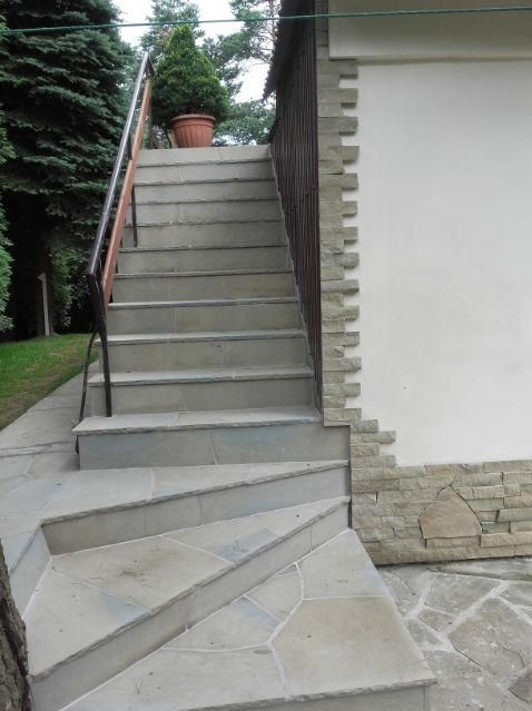 Zewnętrzne schody wyłożone nieregularnymi płytami kamiennymi, prowadzące do domu z elewacją wykończoną dekoracyjnym kamieniem, z donicą z rośliną na górnym podeście.