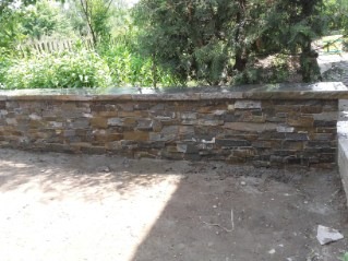 Ściana z kamienia elewacyjnego na zewnątrz budynku, widoczna na tle zieleni ogrodowej, z ciemnym cieniem na ziemi.