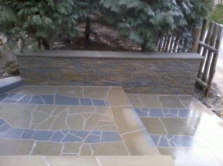 Wykończony taras z mozaikową posadzką w odcieniach beżu i błękitu, prowadzący do kamiennej ścianki oporowej z widocznym fragmentem drewnianego ogrodzenia i iglastego drzewa w tle.