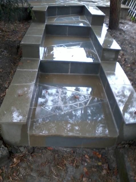 Zewnętrzne schody z szarej kamiennej kostki brukowej, prowadzące w górę po zboczu, z widocznymi refleksami wody na powierzchni. Widoczne liście na ziemi obok schodów.