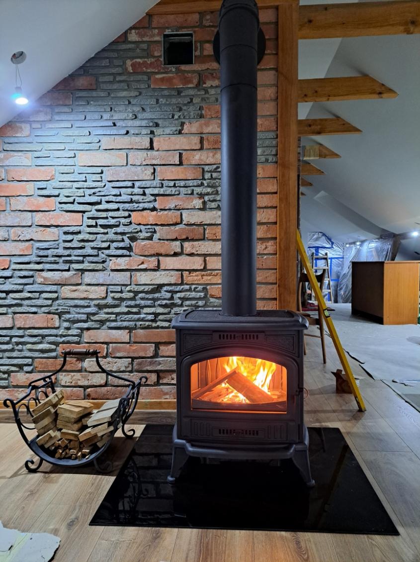 Czarny piec wolnostojący typu koza z palącym się drewnem na tle ściany z cegły i kamienia w trakcie prac wykończeniowych na poddaszu, widoczna drabina i folia ochronna.