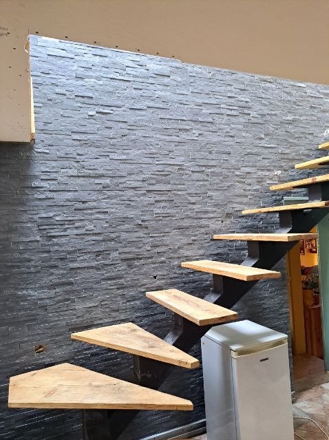 Ściana wykończona szarą okładziną imitującą kamień, z drewnianymi schodami wspartymi na czarnej konstrukcji, obok stoi biała lodówka.