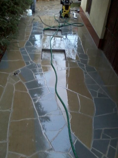 Czyszczenie kamiennej posadzki na zewnątrz budynku za pomocą myjki ciśnieniowej. Widoczne mokre, błyszczące kamienie o nieregularnych kształtach i zielony wąż myjki.