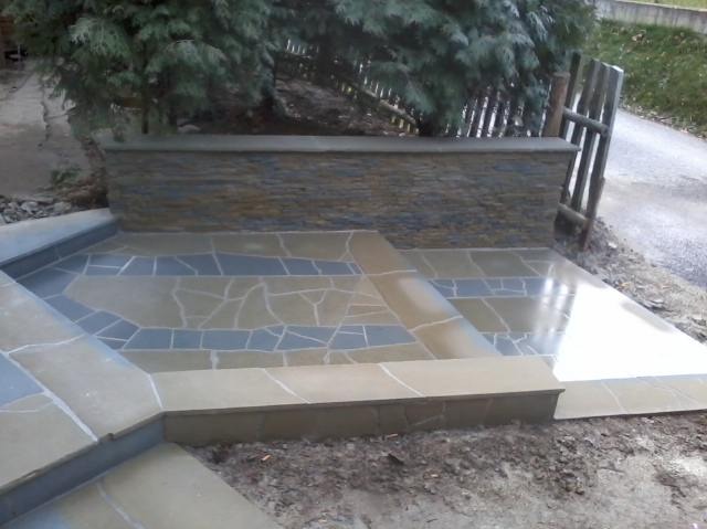 Wykończone schody wejściowe z płyt kamiennych o nieregularnych kształtach w odcieniach beżu i szarości, prowadzące do tarasu z kamienną ścianką, widok z dołu.