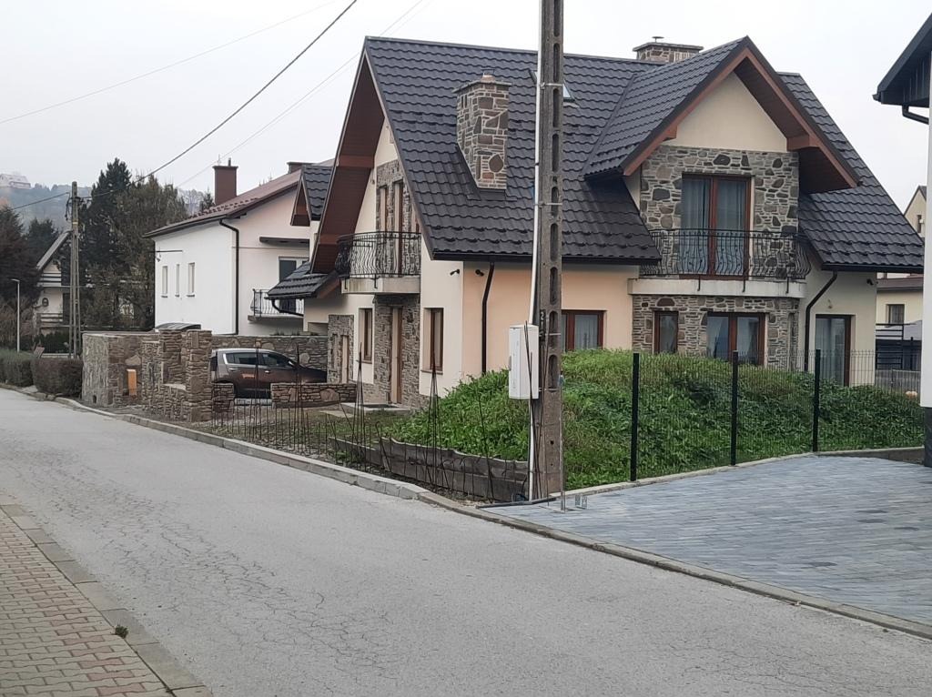 Nowo wybudowany dom jednorodzinny z kamienną elewacją i balkonem, widoczny z ulicy w pochmurny dzień, z fragmentem ogrodzenia i chodnika z kostki brukowej.