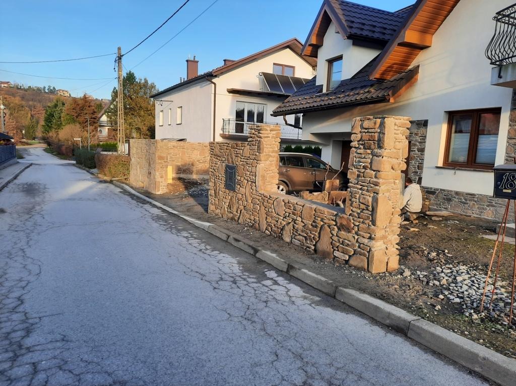 Kamienna zabudowa ogrodzenia posesji z widocznym numerem domu, samochodem na podjeździe i pracującym mężczyzną w tle.