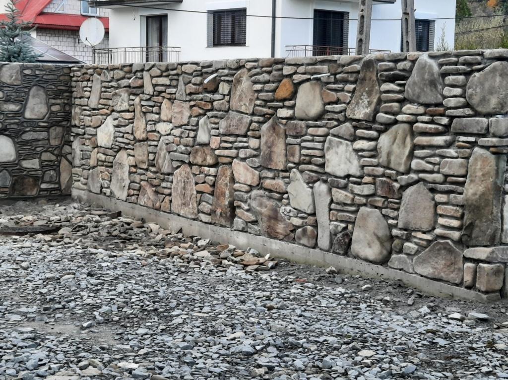 Kamienny mur ogrodzeniowy z nieregularnych, szarych i brązowych kamieni różnej wielkości, z widocznym betonowym fundamentem i gruzem na ziemi.