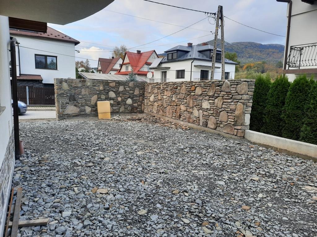 Dwa niskie mury wykończone kamieniem polnym, oddzielające posesję od drogi wysypanej żwirem, z widokiem na okoliczne domy i zalesione wzgórza w tle.