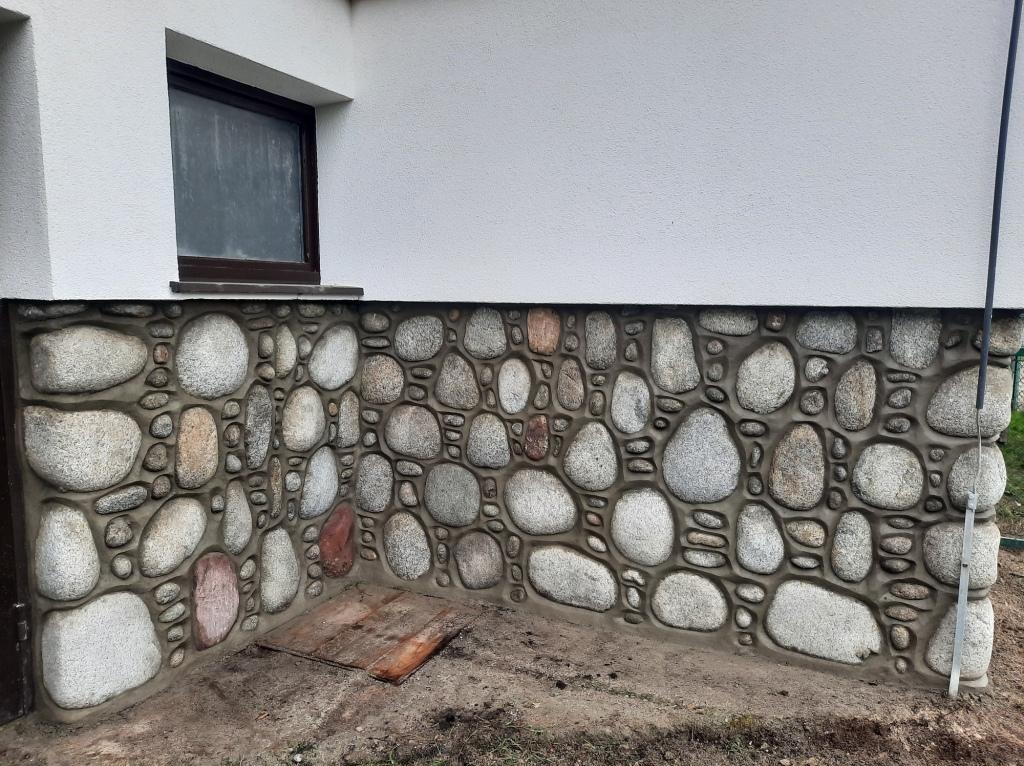 Elewacja budynku z dekoracyjnym wykończeniem z otoczaków osadzonych w zaprawie, widoczny fragment okna i drewniana klapa włazu.