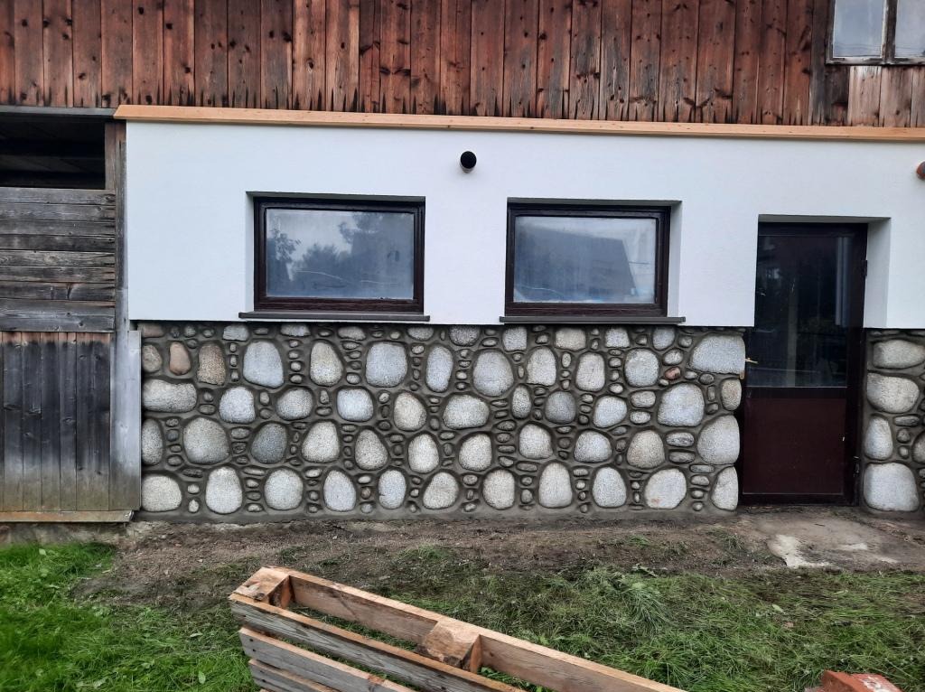 Wykończenie elewacji budynku z wykorzystaniem kamienia naturalnego, widoczne dwa okna z ciemnymi ramami i brązowe drzwi wejściowe, fragment drewnianej palety na trawie.