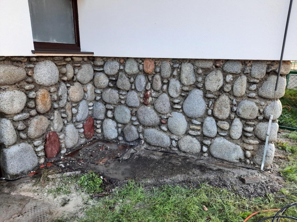 Kamienna podmurówka domu z fragmentami czerwonych kamieni, widoczna dolna część okna i trawa u podstawy ściany.