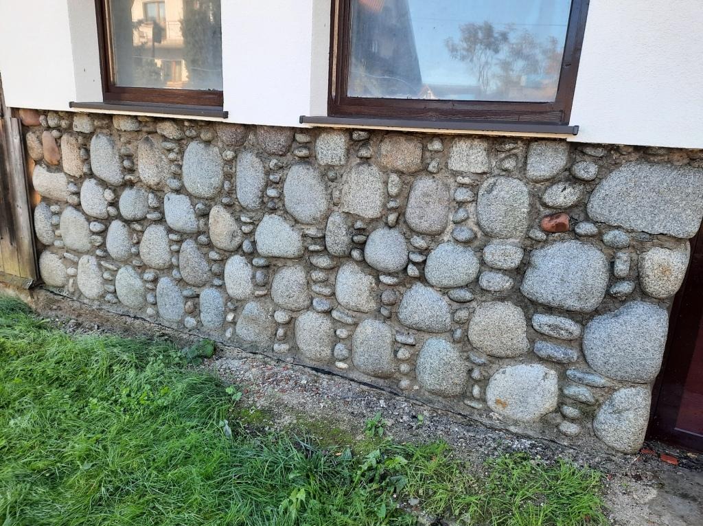 Elewacja budynku wykończona kamieniami rzecznymi, widoczne okna z brązowymi ramami i fragment trawnika.