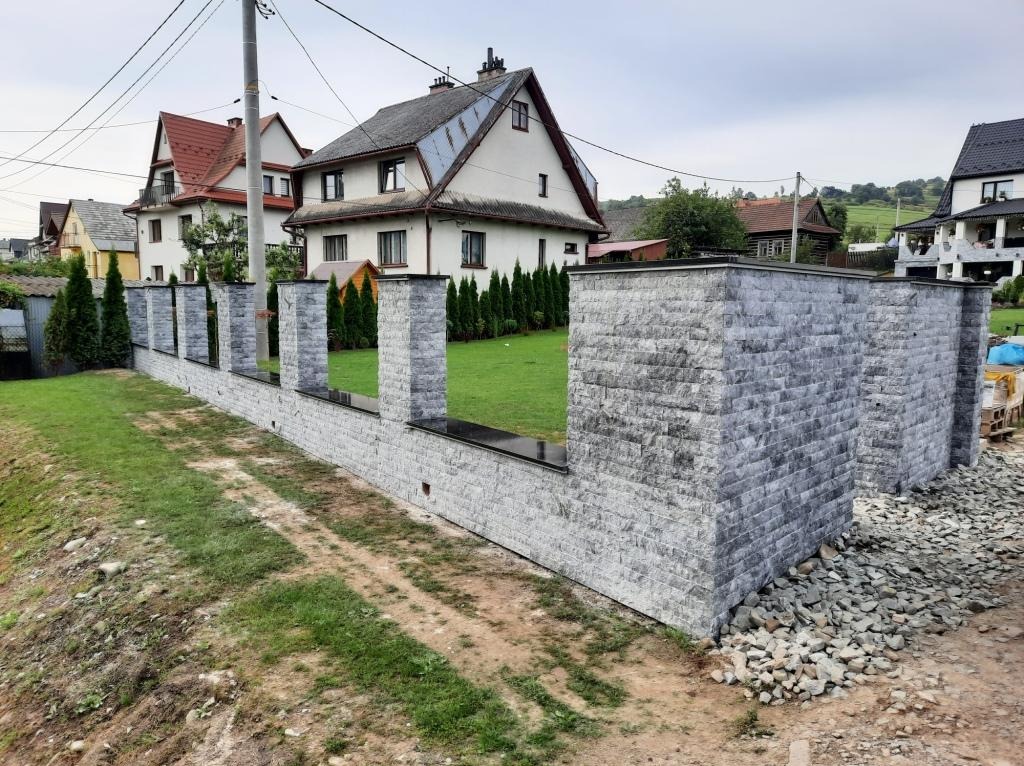 Nowoczesne ogrodzenie z szarej cegły z czarnymi elementami wykończeniowymi w Mszanie Dolnej, widok z perspektywy trawnika, w tle domy i zieleń.