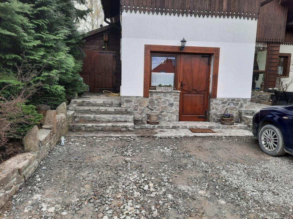 Wejście do domu z drewnianymi drzwiami i kamienną elewacją, schody wyłożone kamieniem, teren wokół wysypany żwirem, widoczny fragment samochodu.