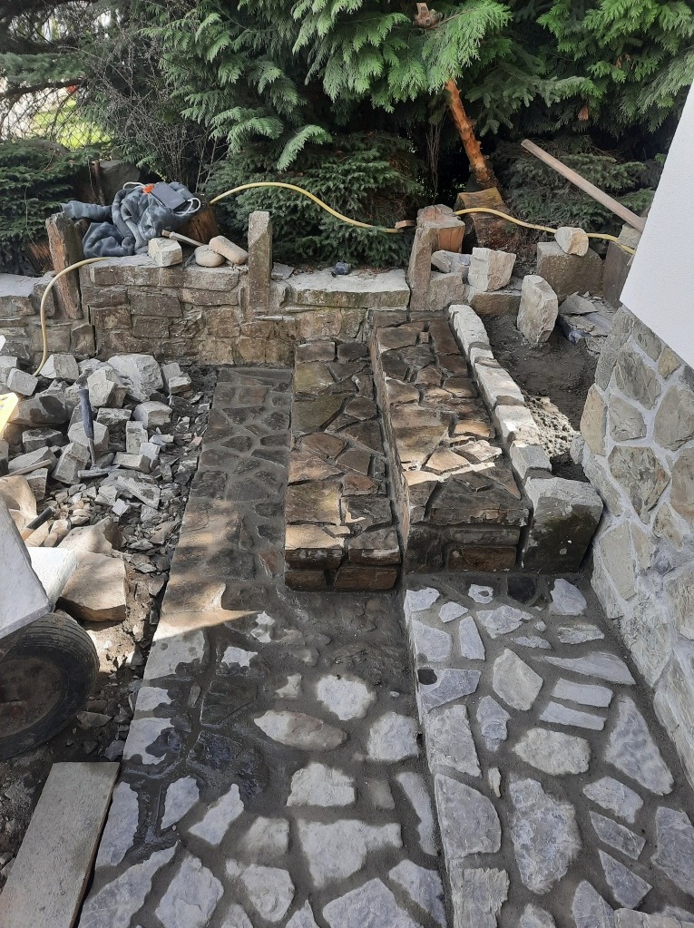 Układanie kamiennej ścieżki w ogrodzie, widoczne fragmenty murku z kamienia, gruz, oraz wąż ogrodowy, etap prac w toku.