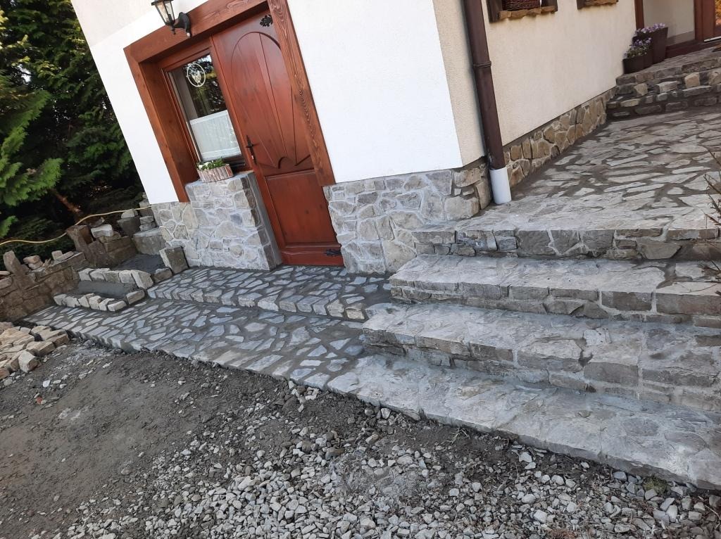 Kamienne schody wejściowe do domu z drewnianymi drzwiami, częściowo wykończone, z widocznym świeżym cementem między kamieniami.