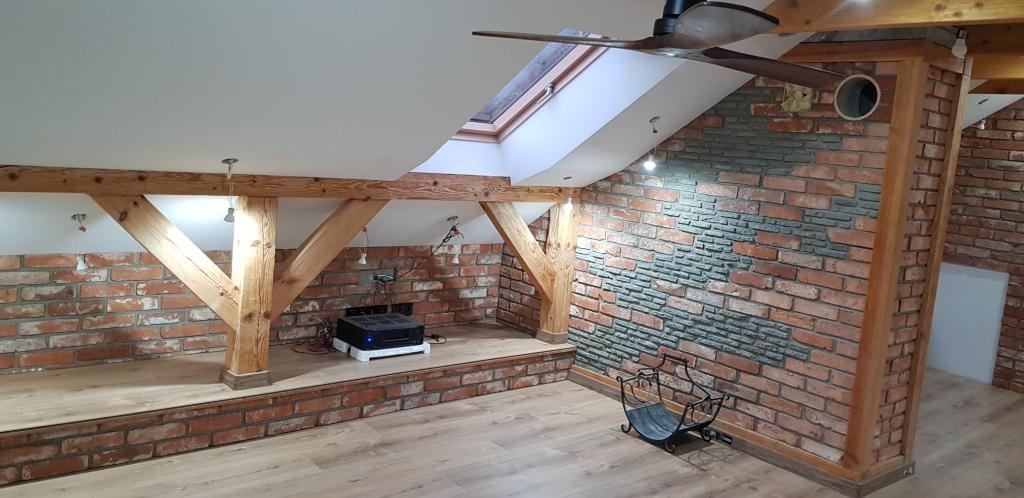Poddasze z ceglaną ścianą, drewnianymi belkami, oknem dachowym i wentylatorem sufitowym, częściowo wykończone szarą okładziną imitującą kamień.