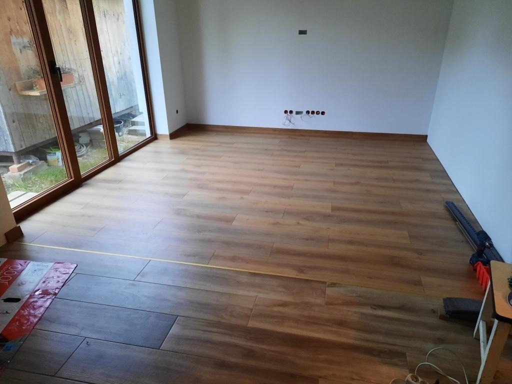 Układanie paneli podłogowych imitujących drewno w jasnym, pustym pokoju z dużym oknem balkonowym, widoczne narzędzia i paczki paneli.