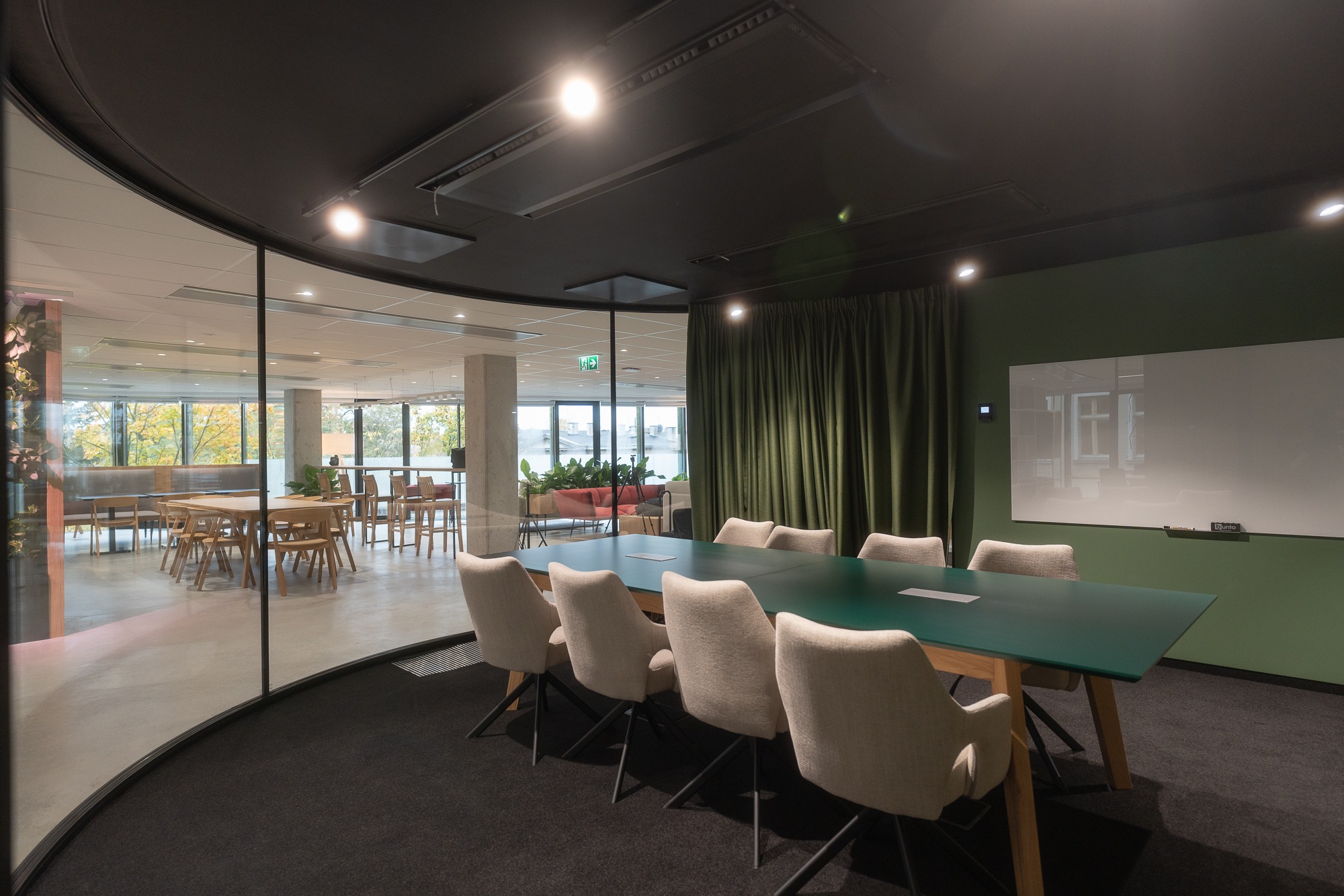 Nowoczesna sala konferencyjna z zielonym stołem, krzesłami w odcieniach beżu, szklaną ścianą oddzielającą przestrzeń biurową z widokiem na zewnątrz, ciemnym sufitem i zieloną ścianą z białą tablicą.