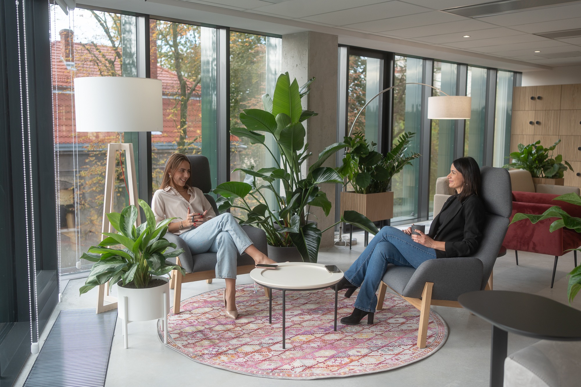 Dwie kobiety w swobodnych strojach siedzą w fotelach w jasnym, nowoczesnym pomieszczeniu z dużymi oknami, roślinami doniczkowymi i okrągłym dywanem z wzorem. Na małym stoliku między nimi leżą...