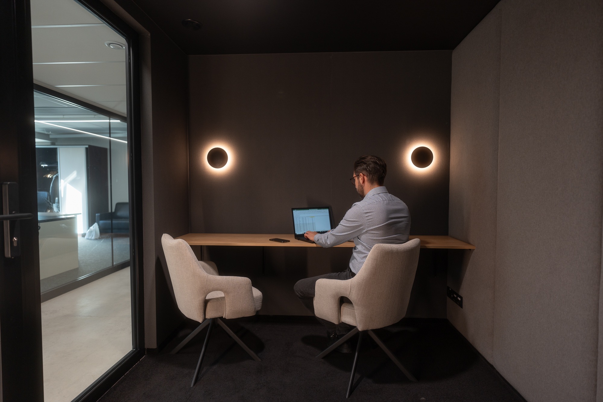 Mężczyzna w okularach pracuje na laptopie w zacisznym, minimalistycznym biurze z dwoma krzesłami, oświetlonym okrągłymi lampami ściennymi, widok z zewnątrz przez szklane drzwi.