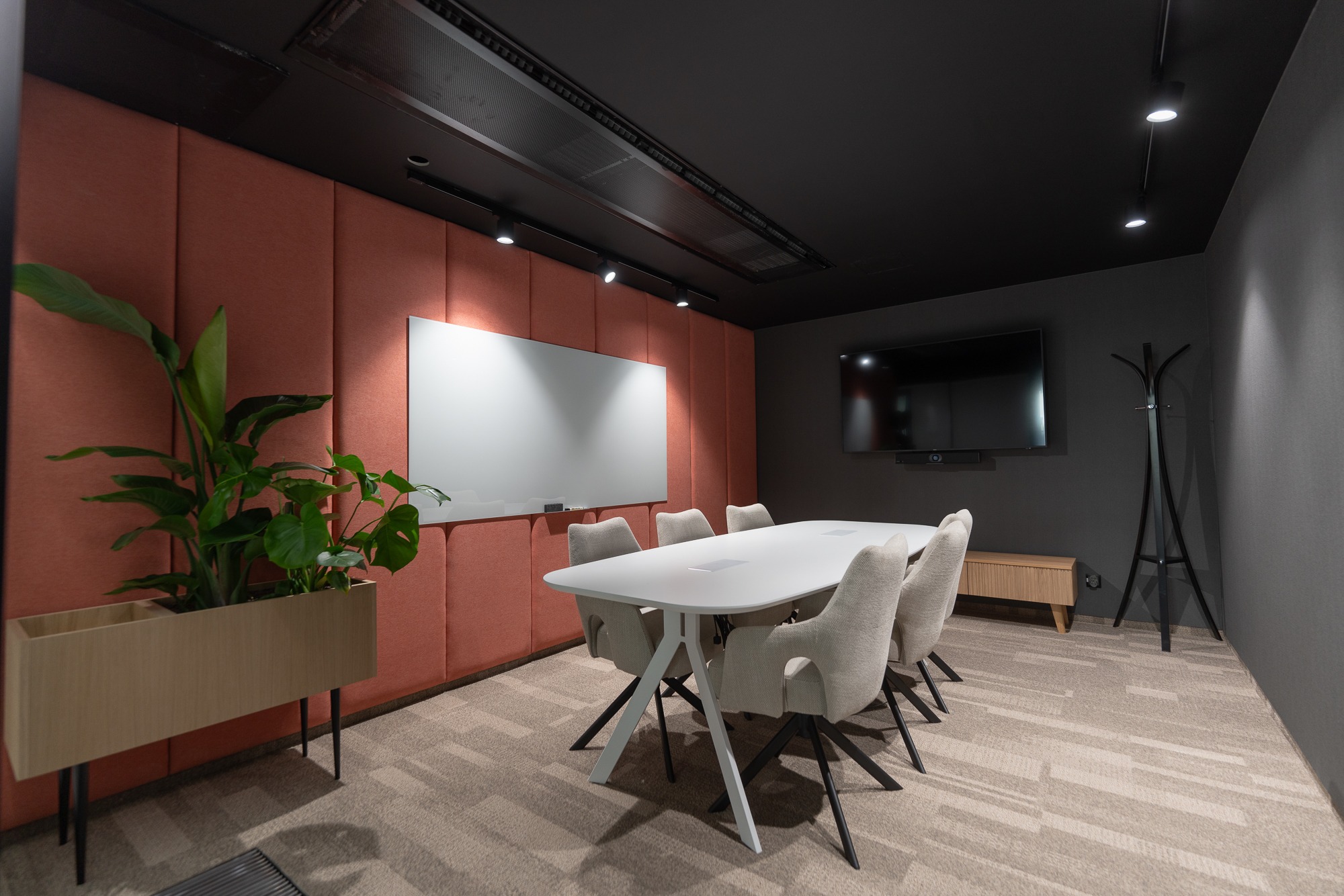 Nowoczesna sala konferencyjna z białym stołem, krzesłami w odcieniach beżu, ścianą w kolorze terakoty, białą tablicą suchościeralną i rośliną w drewnianej donicy.