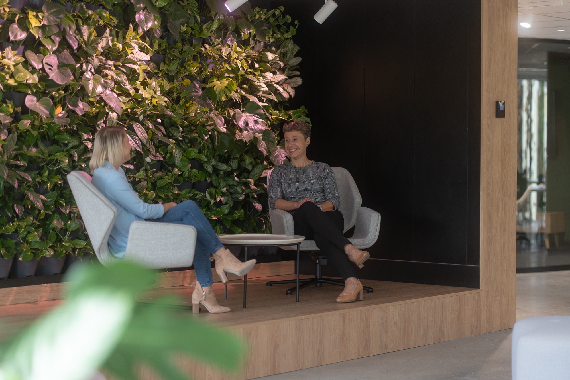 Dwie kobiety siedzą w nowoczesnej przestrzeni biurowej z ścianą z żywych roślin i minimalistycznym wystrojem.