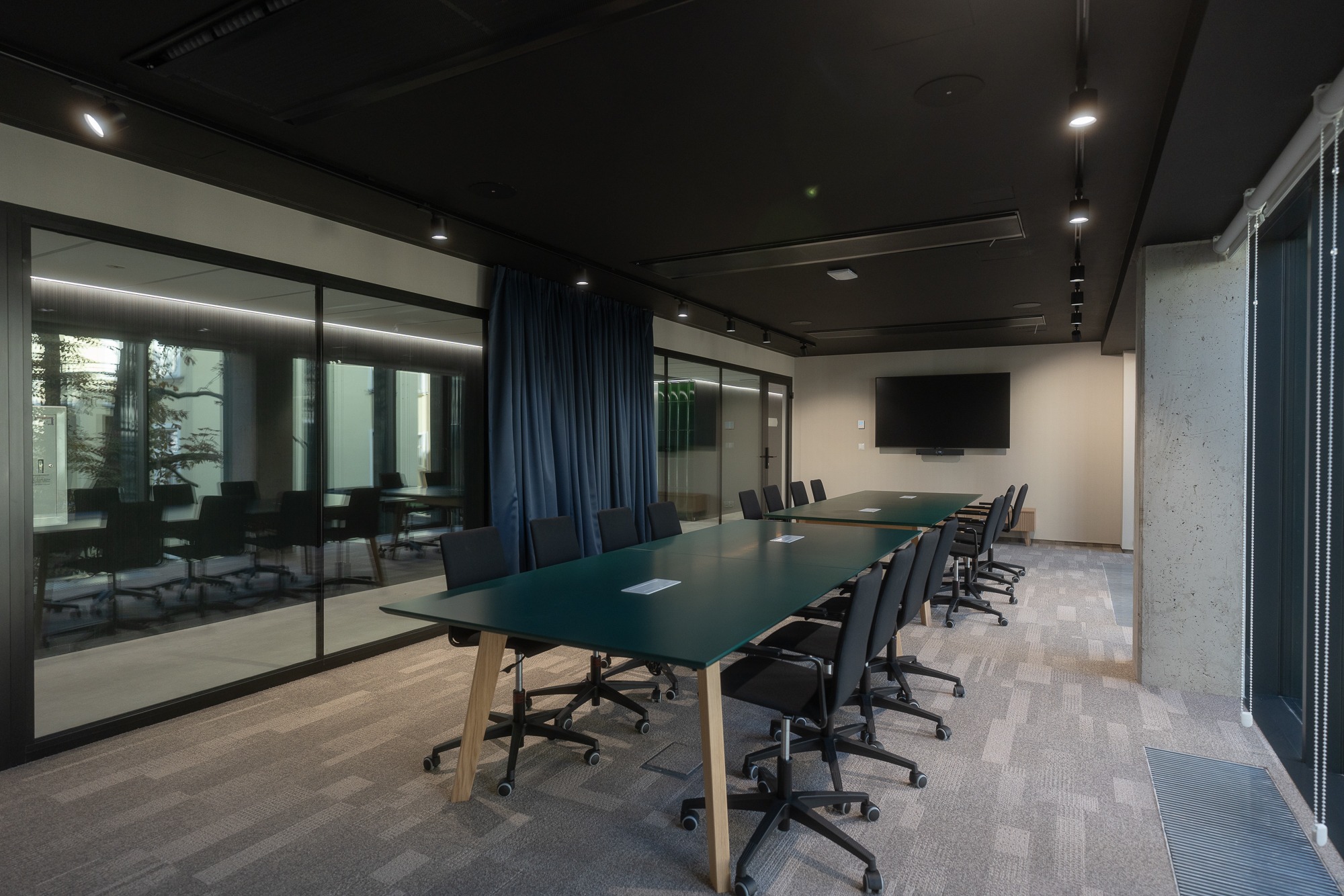Nowoczesna sala konferencyjna z długimi stołami o zielonych blatach i czarnymi krzesłami na kółkach, oświetlona punktowymi lampami sufitowymi, z widokiem na inne pomieszczenia przez szklane ściany...
