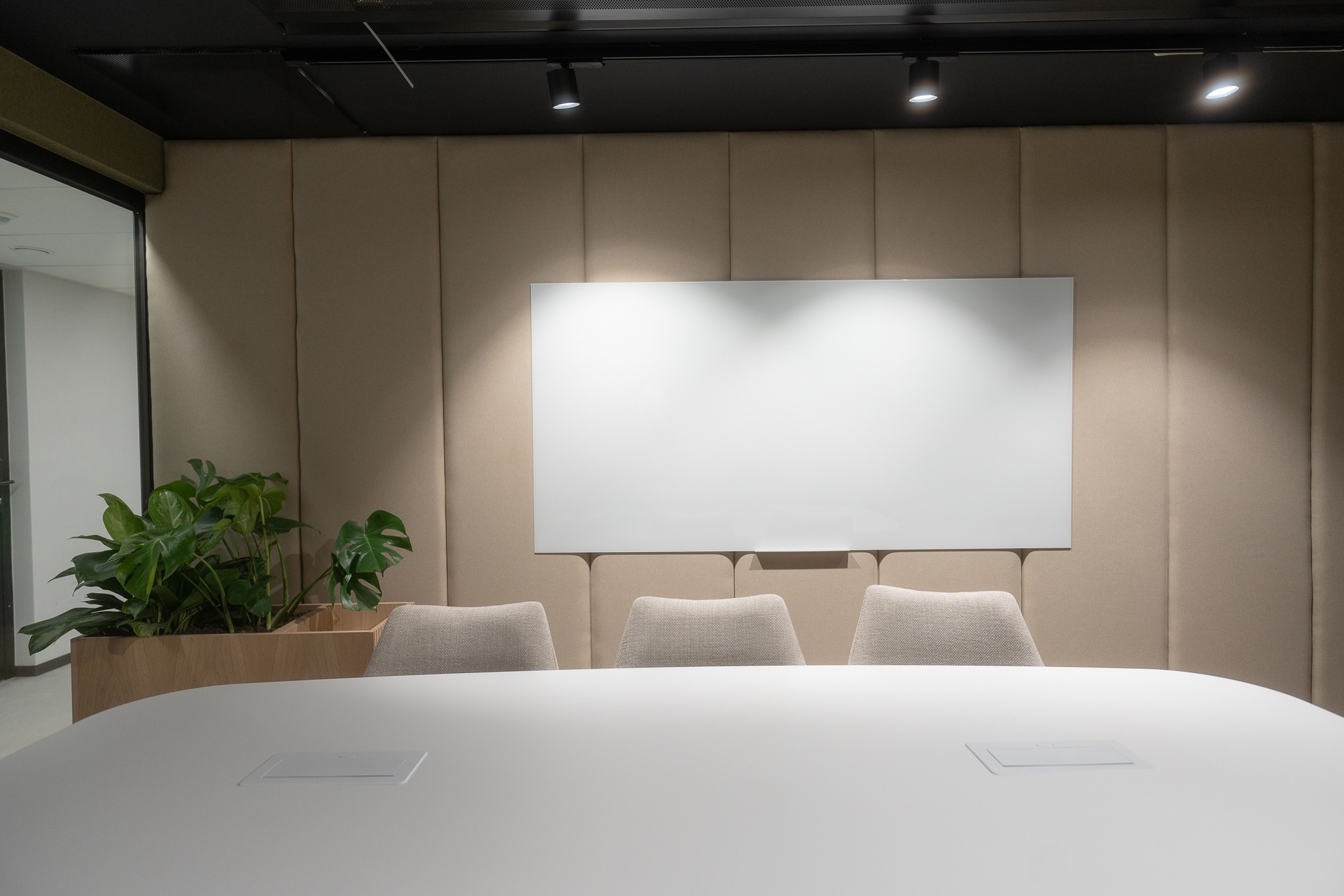 Nowoczesna sala konferencyjna z białym stołem, trzema krzesłami w kolorze beżowym, białą tablicą i rośliną doniczkową w drewnianej skrzynce, oświetlona punktowymi lampami.