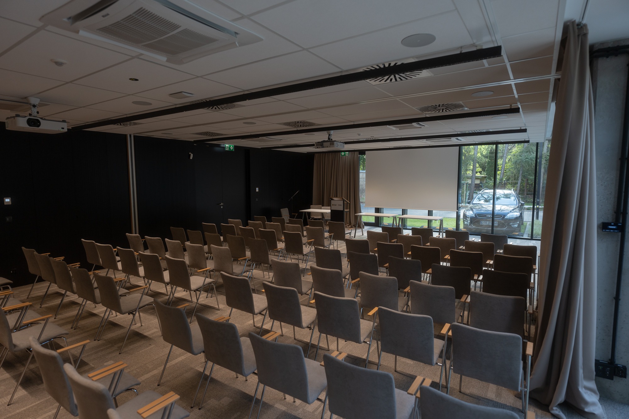 Sala konferencyjna z rzędami krzeseł, projektorem sufitowym i widokiem na samochód za oknem.