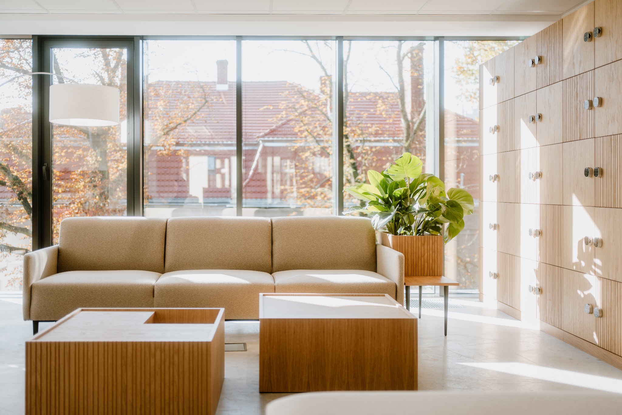 Jasne wnętrze biurowe z beżową sofą, drewnianymi stolikami kawowymi, szafkami i rośliną doniczkową przy dużym oknie z widokiem na budynek z czerwonym dachem.