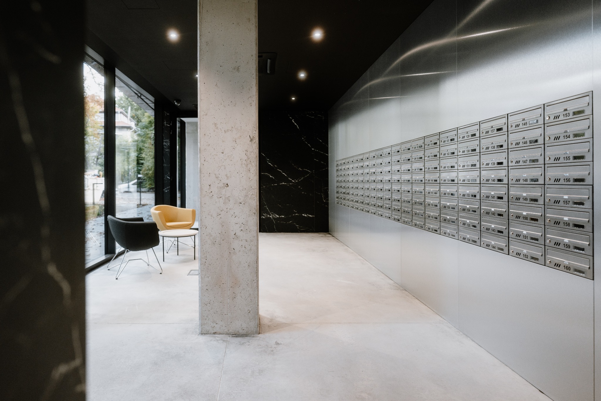 Nowoczesne wnętrze biurowe z rzędem skrytek pocztowych, minimalistycznymi krzesłami i betonowym filarem.