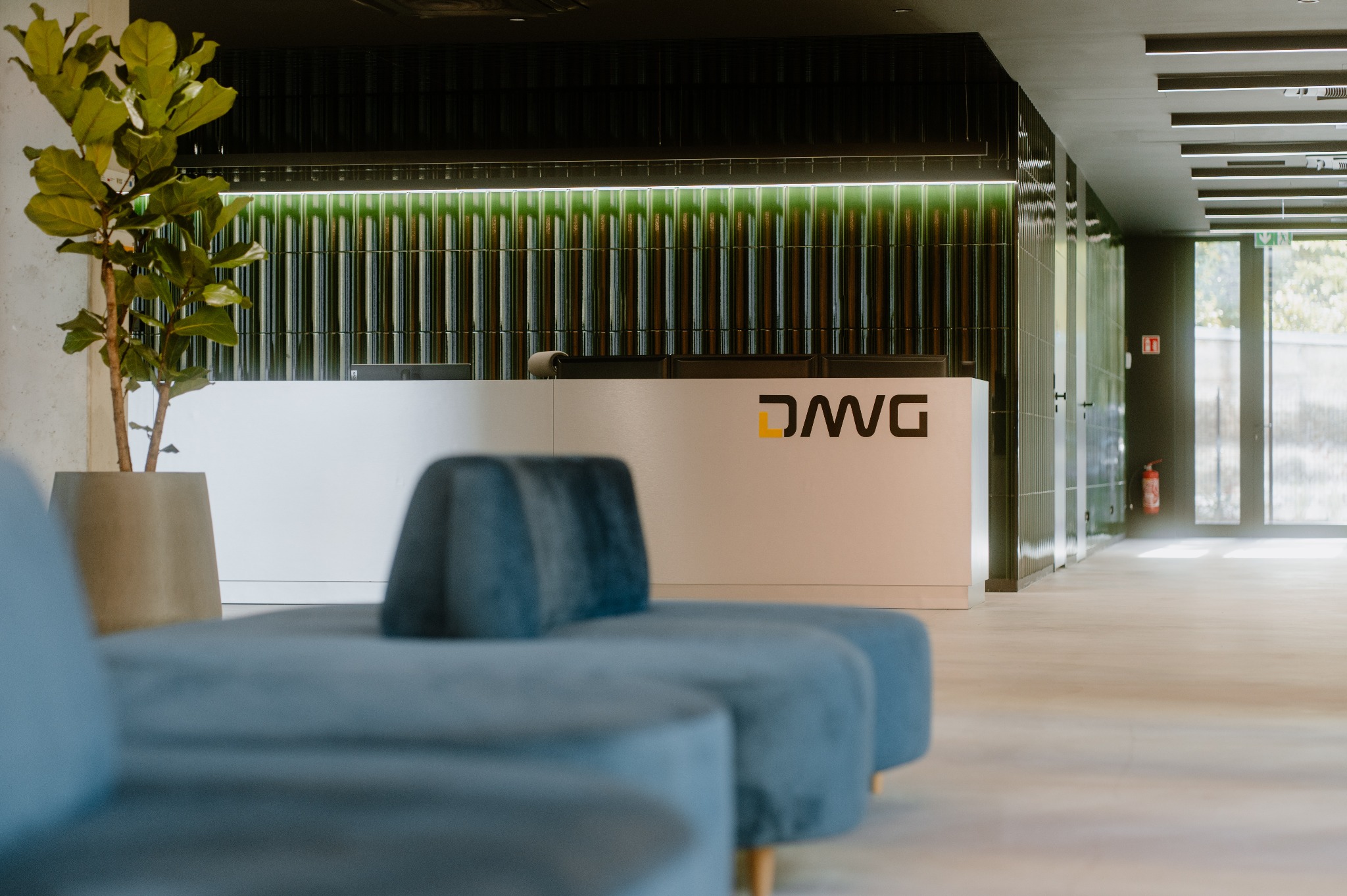 Nowoczesna recepcja z białym biurkiem z logo, zielonym podświetleniem, rośliną w donicy i niebieską sofą w tle, widziana z niskiego kąta.