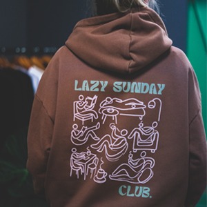 Brązowa bluza z kapturem, na plecach biały nadruk przedstawiający minimalistyczne rysunki osób relaksujących się w różnych pozycjach, z napisem 'LAZY SUNDAY CLUB.'