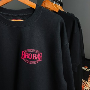 Czarna bluza z okrągłym, różowym nadrukiem 'BBQ BAR BY DI MAURO' w stylu retro, wisząca na drewnianym wieszaku na tle innych ubrań.