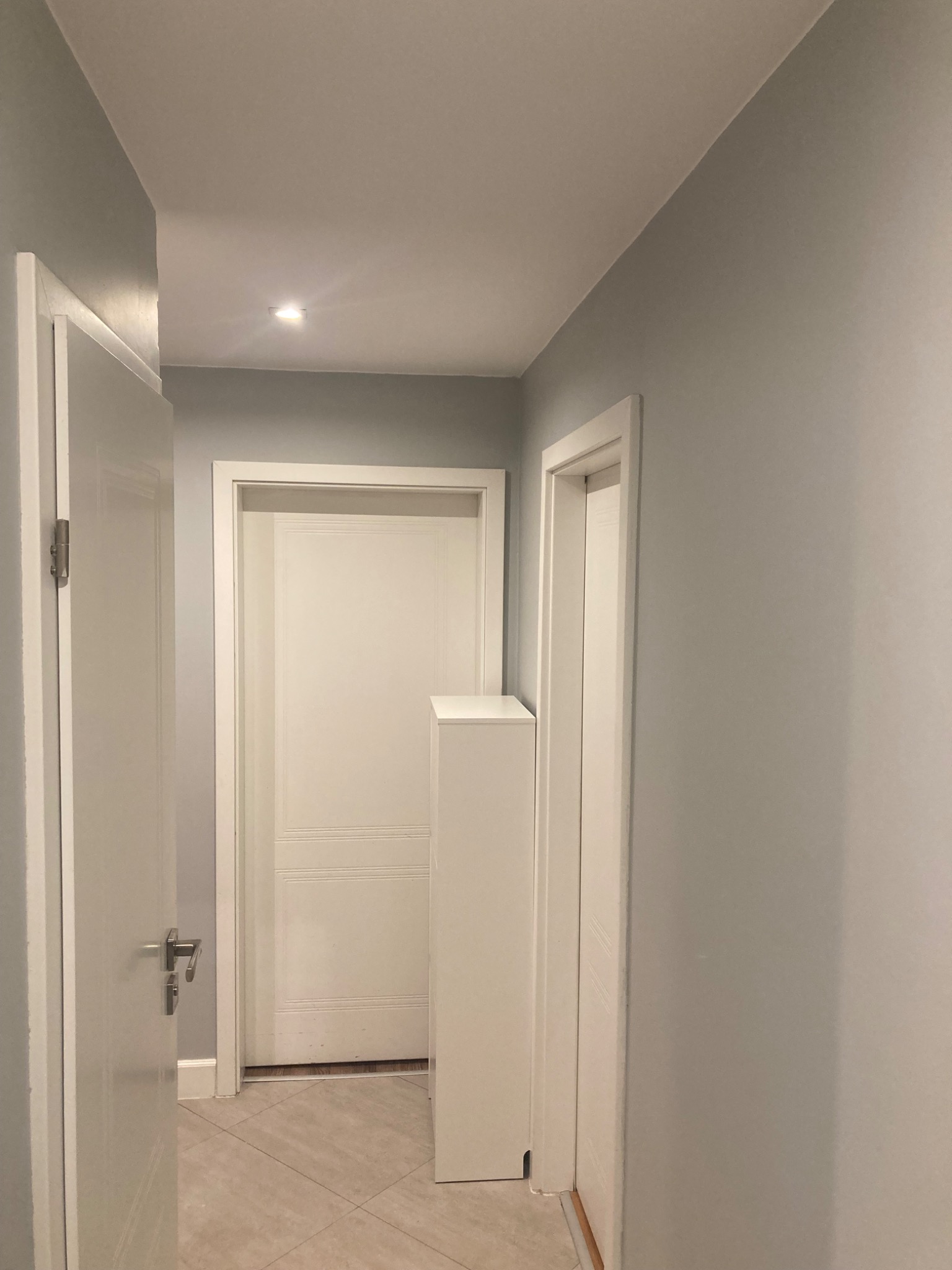 Jasny korytarz po remoncie z pomalowanymi na szaro ścianami, białymi drzwiami i podłogą wyłożoną płytkami w odcieniach beżu.