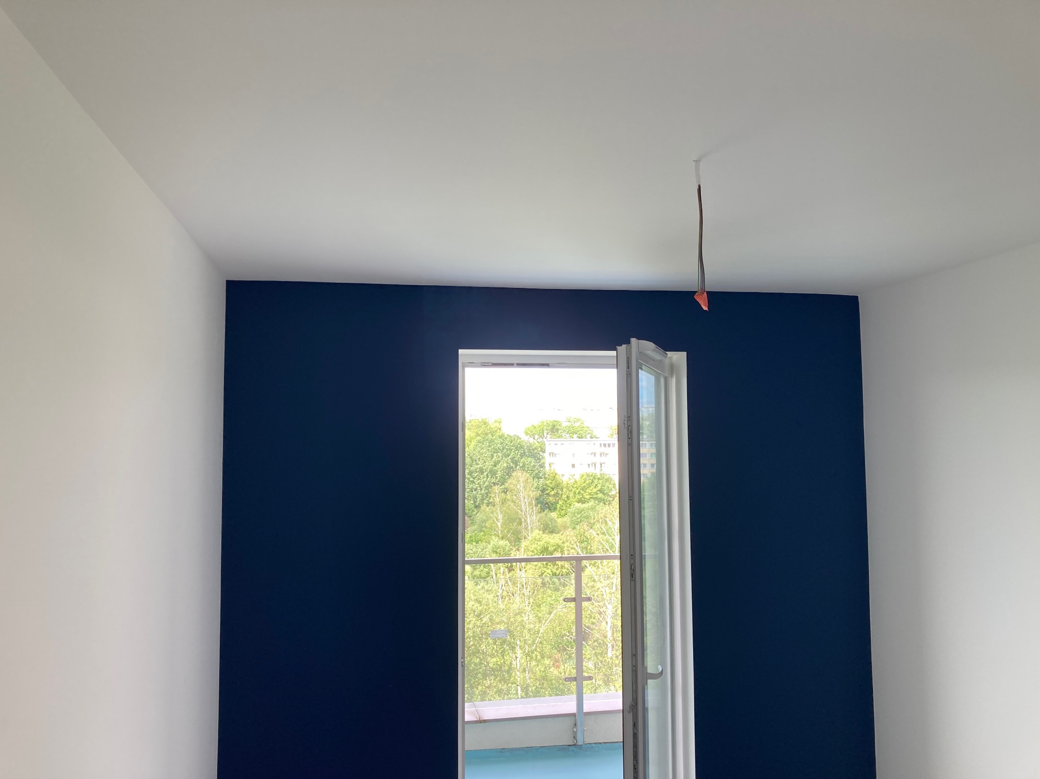 Świeżo pomalowane wnętrze z jedną ścianą w kolorze głębokiego granatu, kontrastującą z białymi ścianami i sufitem, widok przez otwarte okno na zieleń za budynkiem.