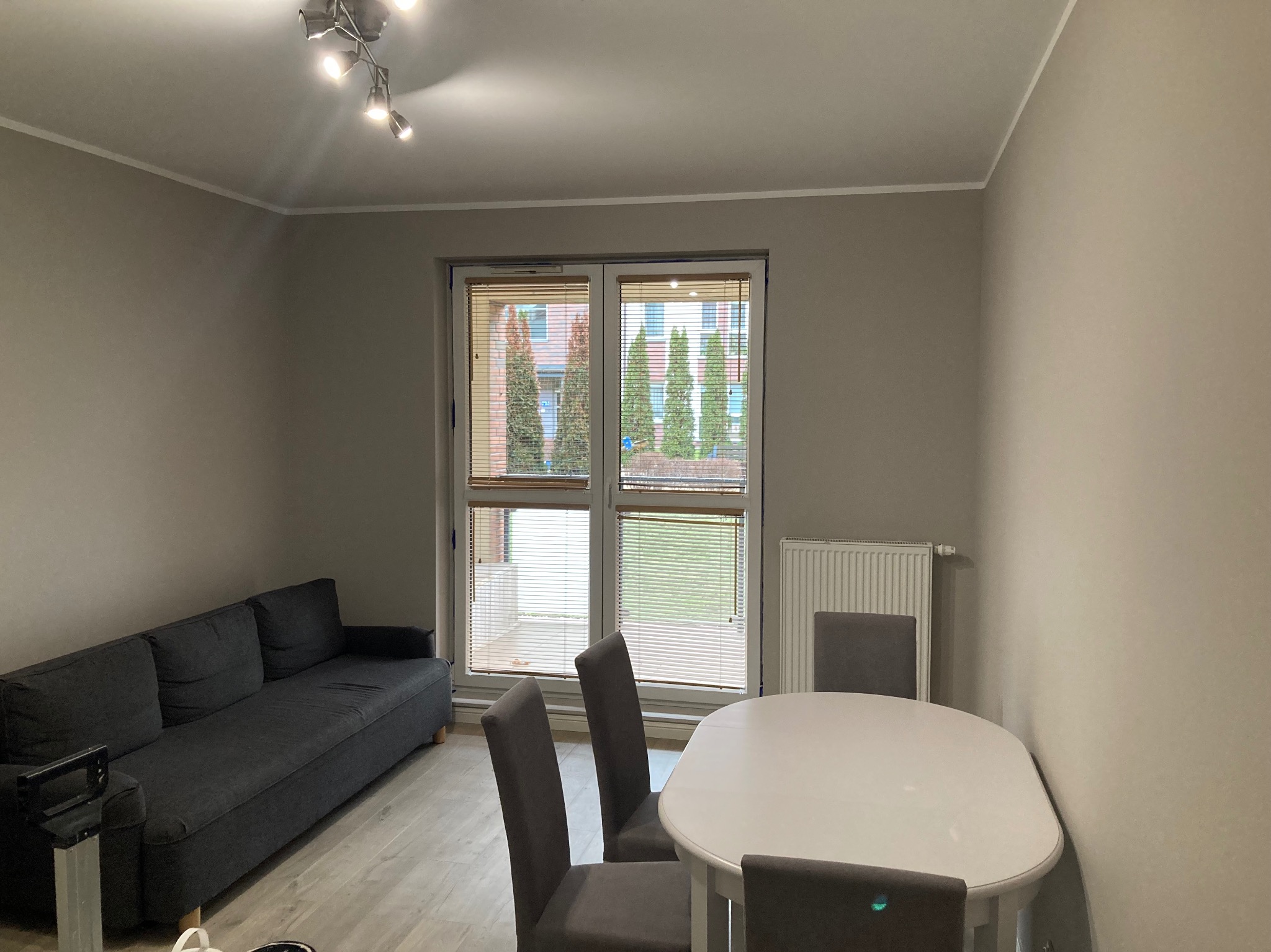 Jasne wnętrze pokoju po odświeżeniu ścian, z szarym narożnikiem, białym stołem i szarymi krzesłami, widok na zewnątrz przez okno z żaluzjami.