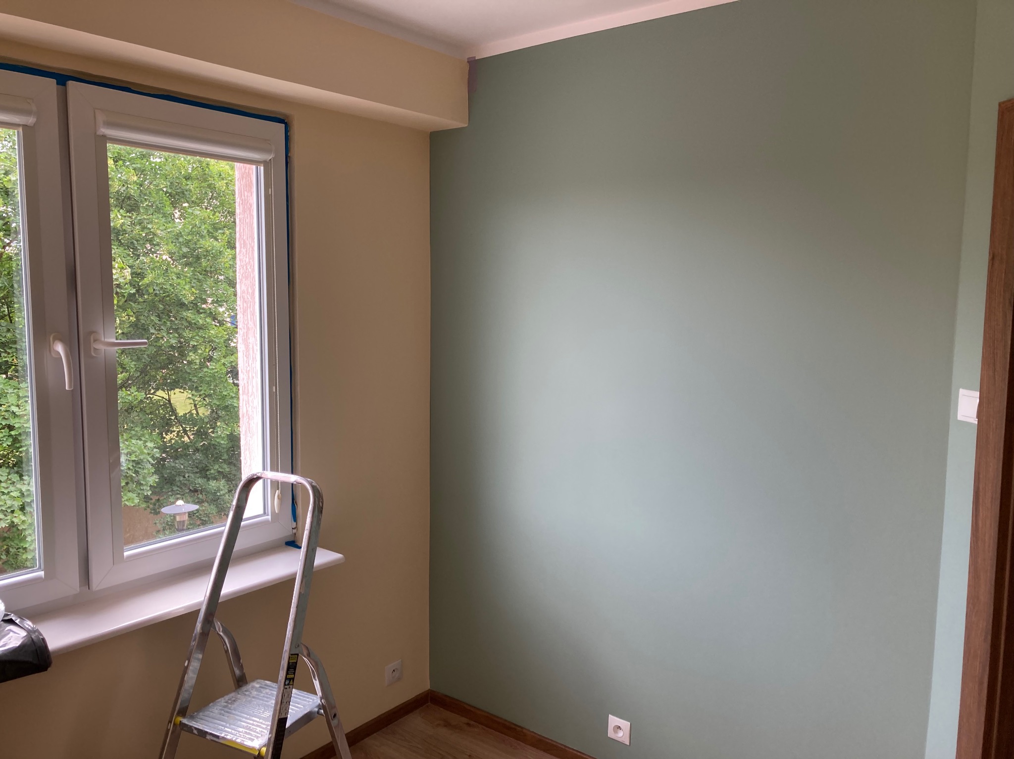 Ściana pomalowana na zielono w Olsztynie, widoczna drabina przy oknie zabezpieczonym taśmą malarską, fragment drugiej ściany w odcieniu beżu.