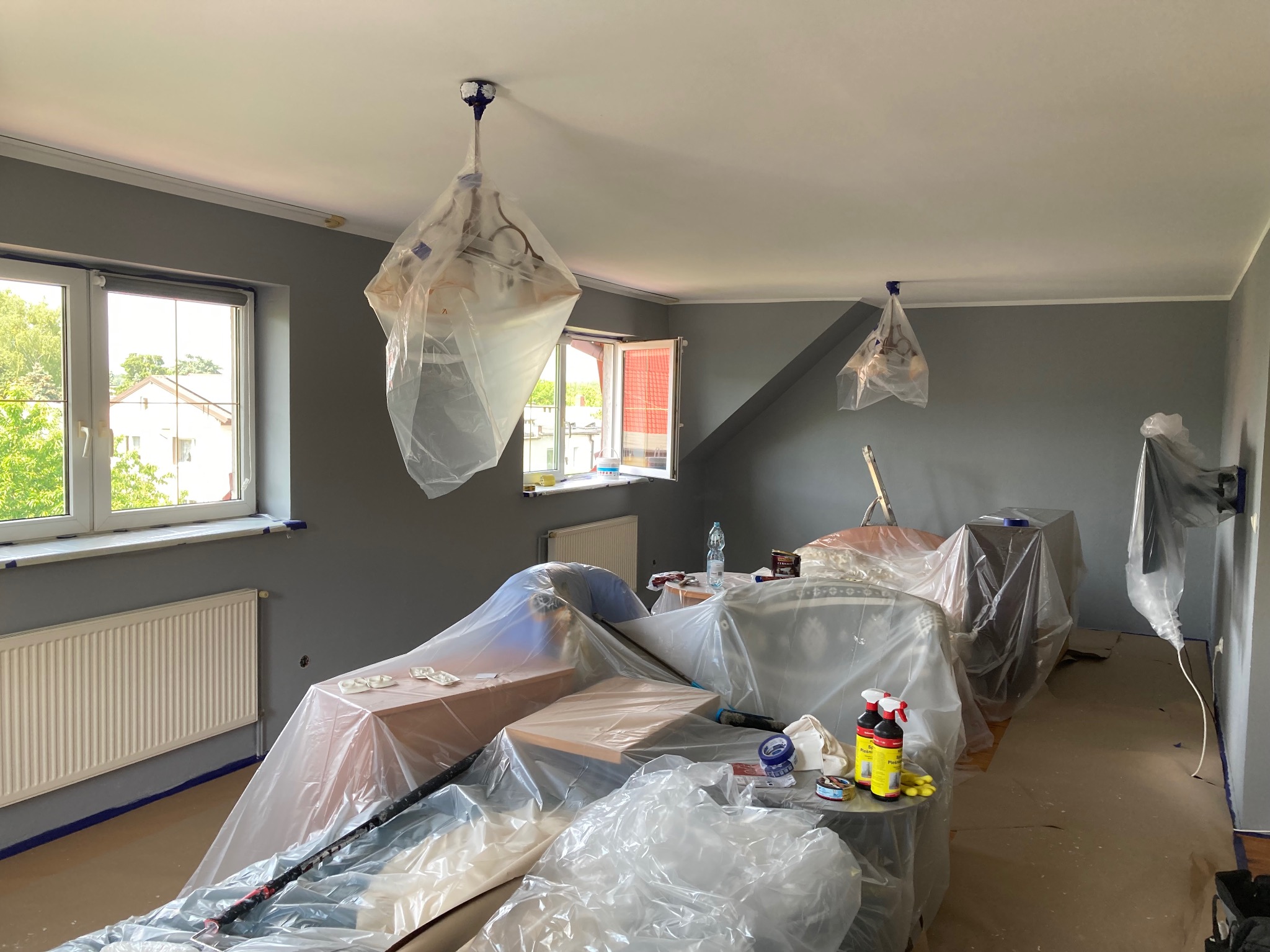 Pokój przed malowaniem: meble zabezpieczone folią, ściany pomalowane na szaro, lampy zakryte folią ochronną, widoczne narzędzia malarskie.