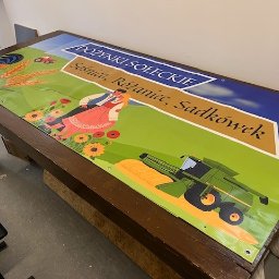 Printio Studio - Baner reklamowy na drewnianym stole, z grafiką dożynek Soleckich: kogut, traktor, para taneczna, kombajn. Żywe kolory, widoczne oczka mocujące.
