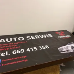 Czarny baner reklamowy auto serwisu z białymi napisami, logotypem i grafiką samochodu, leżący na brązowym stole. Widoczne media społecznościowe i numer telefonu.
