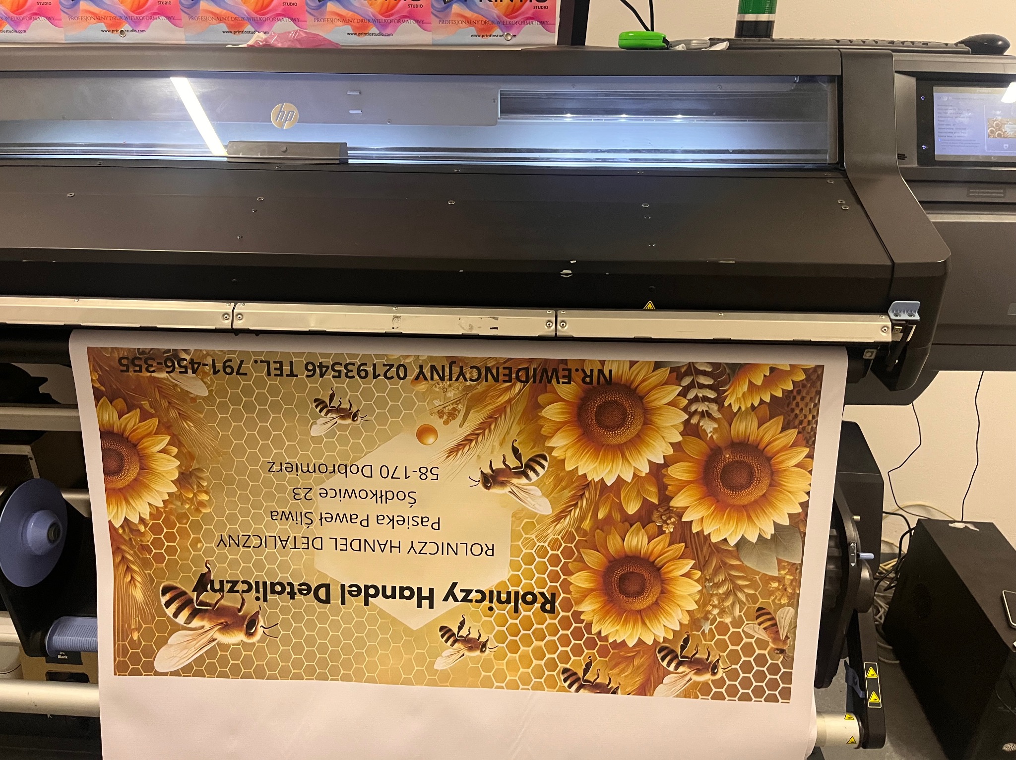 Drukarka wielkoformatowa HP drukuje baner reklamowy z motywem plastra miodu, słoneczników i pszczół, zawierający dane adresowe i kontaktowe pasieki.