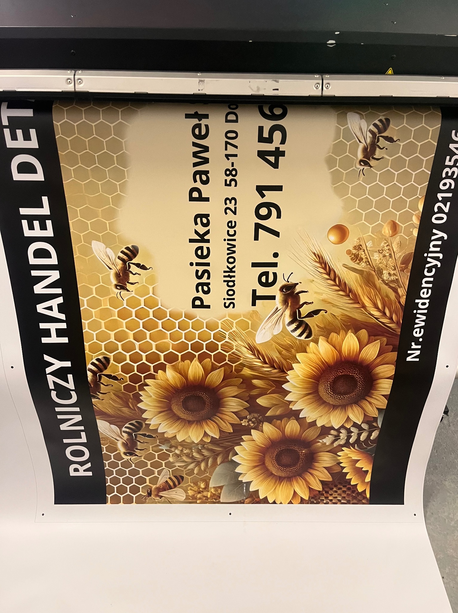 Wydruk wielkoformatowy przedstawiający projekt graficzny dla pasieki z motywem plastra miodu, słoneczników i pszczół, zawierający dane kontaktowe: nazwę firmy, adres i numer telefonu.