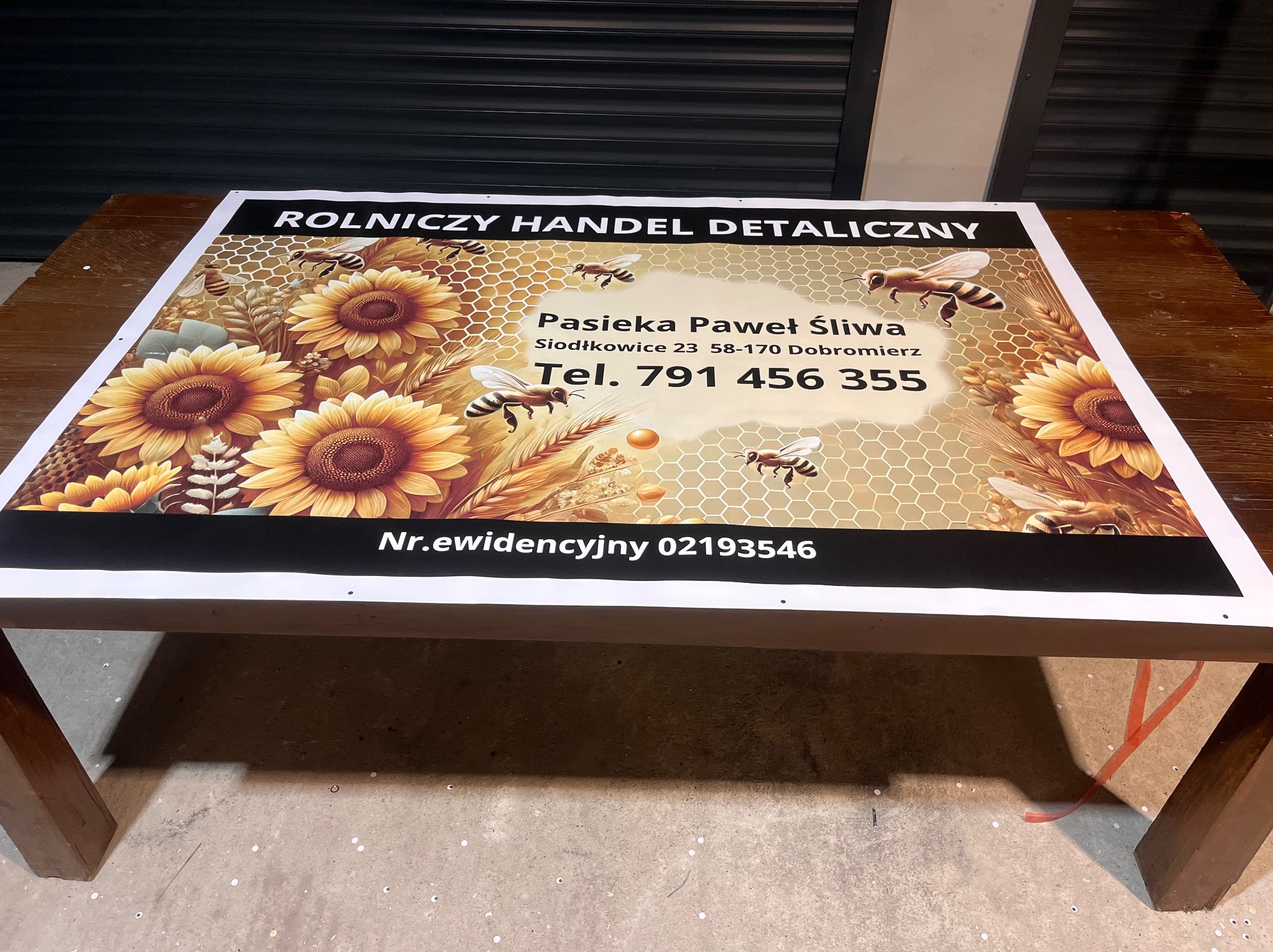 Duży baner reklamowy z motywem plastra miodu, słoneczników i pszczół, z danymi kontaktowymi pasieki Pawła Śliwy, leżący na drewnianym stole.
