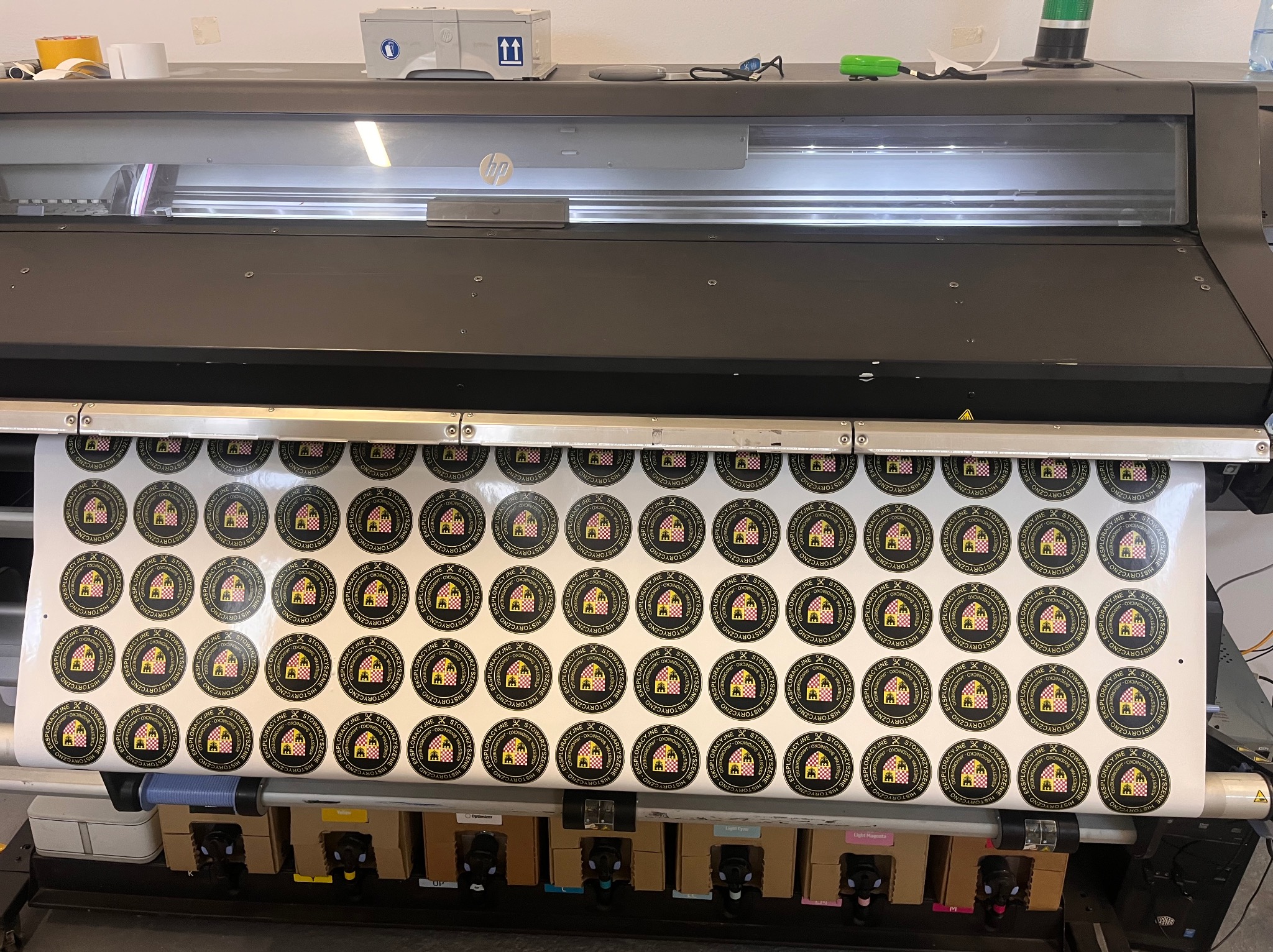 Wielkoformatowa drukarka HP drukująca arkusz okrągłych naklejek z czarno-złotym logo przedstawiającym budynek w kratkę.